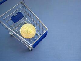 Close-up of a golden bitcoin in the blue metal shopping cart on blue background with copy space for text photo