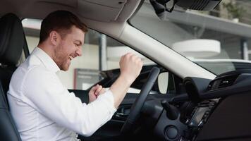 contento hombre se sienta en nuevo coche en tienda concesión y celebrar compra de nuevo vehículo. comprar coche video