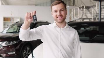 Portrait of happy adult successful man posing in auto showroom buying new automobile. Positive male smiling for camera and demonstrating keys while standing near new vehicle in showroom video