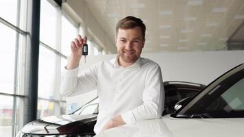 retrato de contento adulto exitoso hombre posando en auto sala de exposición comprando nuevo automóvil. positivo masculino sonriente para cámara y demostrando llaves mientras en pie cerca nuevo vehículo en sala de exposición video