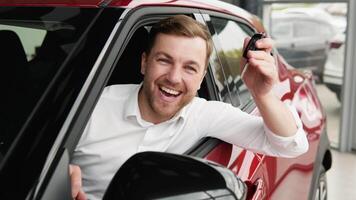 contento hombre se sienta en nuevo coche en tienda concesión y celebrar compra de nuevo vehículo. el hombre con llaves muestra emociones de felicidad mientras conducción en su nuevo coche video