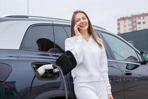 Young woman using a smartphone to pay for electric car at the charging station. Recharging battery from charging station. photo