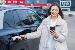 Young beautiful woman traveling by electric car having stop at charging station. Electric car concept. Green travelling. photo