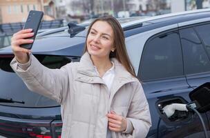 Young woman using a smartphone to pay for electric car at the charging station. Recharging battery from charging station. photo