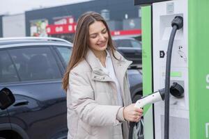 joven mujer utilizando un teléfono inteligente a pagar para eléctrico coche a el cargando estación. recarga batería desde cargando estación. foto