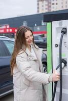 joven mujer utilizando un teléfono inteligente a pagar para eléctrico coche a el cargando estación. recarga batería desde cargando estación. foto