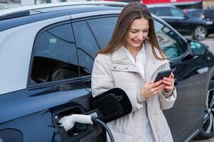 joven mujer cargando su eléctrico coche a un cargando estación en el ciudad. eco combustible concepto. el concepto de ambientalmente simpático transporte. recarga batería desde cargando estación. foto