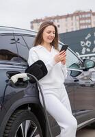 Young woman using a smartphone to pay for electric car at the charging station. Recharging battery from charging station. photo