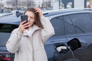 Young woman using a smartphone to pay for electric car at the charging station. Recharging battery from charging station. photo