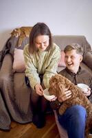 couple drinking from cups on couch in living room, ginger cat sleeping on couch, cockapoo also drinking from mom's cup photo