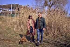 a young stylish couple walking with their cockapoo dog near the river, an unfinished parking lot in the background photo