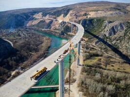 Aerial drone view of the construction of a high concrete bridge over the river. Highway bridge under construction. Several high crane machines and workers on the construction site. photo