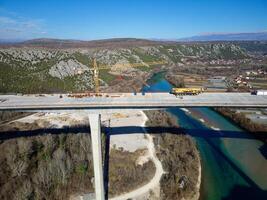 Aerial drone view of the construction of a high concrete bridge over the river. Highway bridge under construction. Several high crane machines and workers on the construction site. photo