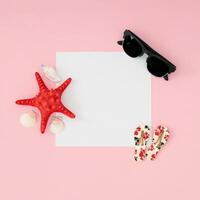 Trendy summer layout made with sunglasses, red starfish, sea shells, floral slippers and paper card note copy space on pastel pink background. Minimal concept. Summer aesthetic. Flat lay. photo