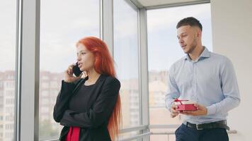 un hombre da un regalo a un mujer con rojo cabello. un mujer quien habla en el teléfono recibe un regalo desde su marido y se regocija a el regalo recibido en el oficina. video