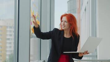 Büro Arbeit mit Laptop und Aufkleber. jung rothaarig Frau hält ein Laptop im ihr Hand und nimmt ein Aufkleber von das Fenster und untersucht Es. Geschäft Dame im das Büro beim arbeiten. video