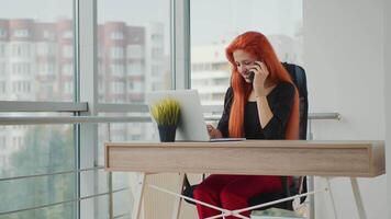 een jong geslaagd bedrijf dame en bedrijf manager, werken in haar kantoor Bij een computer en lost op gevallen Aan een mobiel telefoon. jong geslaagd vrouw met rood haar- in de kantoor. video