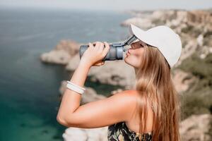 mujer viaje mar. contento turista bebida agua en caliente verano día. mujer viajero mira a el borde de el acantilado en el mar bahía de montañas, compartiendo viaje aventuras viaje foto