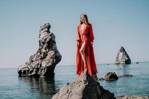 Woman travel sea. Young Happy woman in a long red dress posing on a beach near the sea on background of volcanic rocks, like in Iceland, sharing travel adventure journey photo