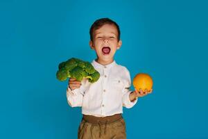 A studio shot of a boy holding fresh broccoli and an orange on a blue background. The concept of healthy baby food. photo