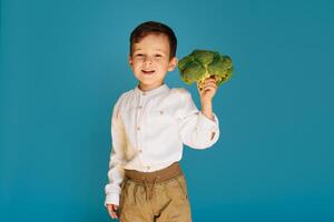 un estudio Disparo de un chico participación Fresco brócoli en un azul antecedentes. el concepto de sano bebé alimento. foto