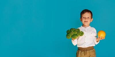 A studio shot of a boy holding fresh broccoli and an orange on a blue background with a copy of the space. The concept of healthy baby food. photo