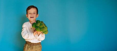 A studio shot of a smiling boy holding fresh broccoli on a blue background with a copy of the space. The concept of healthy baby food. photo