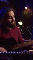 Vertical Video Songwriter producing tunes on acoustic guitar in his personal studio, using a microphone to sing the chords. Audio engineer works with soundboard and equalizer to compose new music. Camera B.