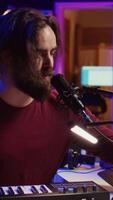 verticale vidéo artiste en chantant une chanson à microphone et en jouant électronique piano à créer la musique dans Accueil studio. auteur compositeur enregistrement midi manette des sons, ajustement le volume avec pore ampli boutons. caméra b. video