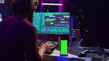 Sound engineer working on mixing and mastering techniques in his home studio, using phone with greenscreen display while he is recording audio. Artist operating soundboard and amplifier. Camera B. video