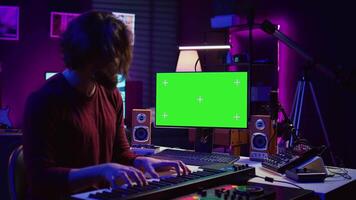 Artist learning to play at electronic piano keyboard in home studio, watching tutorial online with greenscreen display. Music producer practices playing on midi controller synthesizer. Camera B. video