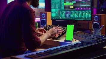 Sound engineer working on mixing and mastering techniques in his home studio, using phone with greenscreen display while he is recording audio. Artist operating soundboard and amplifier. Camera A. video