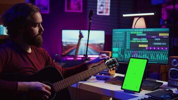 Audio engineer practices tuning his acoustic guitar in home studio, watching a video tutorial online via tablet with greenscreen. Musician learning to play instrument and create music. Camera B.