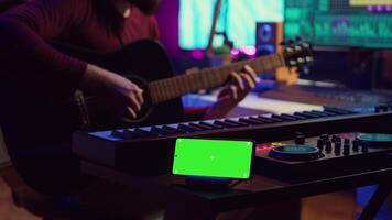 Songwriter playing acoustic guitar in home studio with greenscreen display running on smartphone app. Musician practices singing on musical instrument with strings, isolated screen. Camera A. video