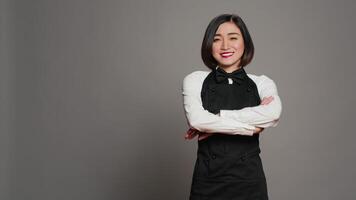 Restaurant Hostess posieren mit Vertrauen im Studio, Stehen mit Waffen gekreuzt Über grau Hintergrund. asiatisch Kellnerin Barista mit Gourmet Portion Sachverstand lächelnd auf Kamera. Kamera a. video