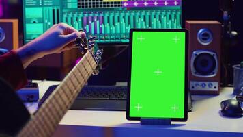 Audio engineer practices tuning his acoustic guitar in home studio, watching a video tutorial online via tablet with greenscreen. Musician learning to play instrument and create music. Camera A.