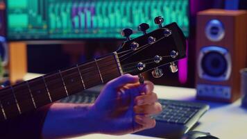 Musical performer tuning his guitar by twisting the knobs, preparing to play acoustic instrument in home studio. Artist recording sounds to create new track, using equalizer mixing gear. Camera A. video