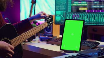 Musical producer learning to tune his guitar before playing, watching lesson tutorial video by using greenscreen display on tablet. Young artist tuning strings on instrument, practicing. Camera A.