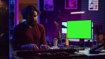 Artist learning to play at electronic piano keyboard in home studio, watching tutorial online with greenscreen display. Music producer practices playing on midi controller synthesizer. Camera A. video