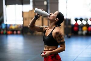 Tired strong mature sportswoman drinking water from a flask indoors photo