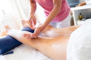 Crop masseuse doing body massage with oily cream on leg of female client photo