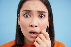 Portrait of shocked korean girl, gasping and looking worried, frowning, looks scared and anxious, stands over blue background photo