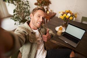 Man taking selfie with thumbs up, sitting in cafe with his laptop and smiling at camera, blogger posting on social media app photo