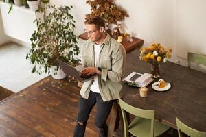 Handsome manager, man in glasses sits on table, holds laptop and types, sends message, works on project, drinks coffee, spends time at co-working space or an office photo