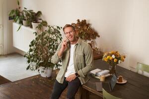 Portrait of handsome happy man in an office, guy sits in coworking space, looking at camera, smiling and laughing, talking on mobile phone, using smartphone, answer a call from client photo