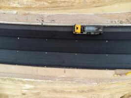 yellow truck pouring bitumen over a new road. Aerial drone shot. Road construction in progress on slope nature canyon. Infrastructure development and logistics. photo