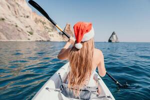 Woman in kayak back view. Happy young woman in Santa hat floating in kayak on calm sea. Summer holiday vacation and cheerful female people relaxing having fun on the boat. photo