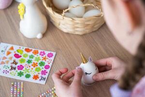 A cute girl with pink bunny ears makes an Easter craft - decorates an egg in the form of a unicorn with rhinestones, horn, flowers in the interior of a house with plants. photo