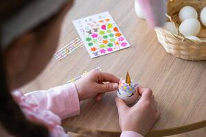 A cute girl with pink bunny ears makes an Easter craft - decorates an egg in the form of a unicorn with rhinestones, horn, flowers in the interior of a house with plants. photo