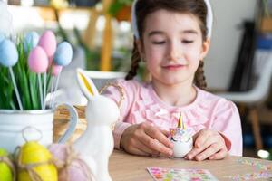 un linda niña con rosado conejito orejas hace un Pascua de Resurrección arte - decora un huevo en el formar de un unicornio con pedrería, bocina, flores en el interior de un casa con plantas. foto